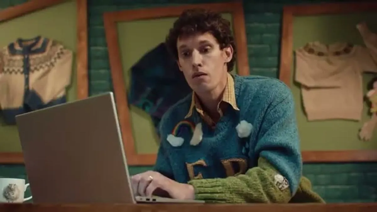 A man with curly brown hair and a blue and green sweater sits at a laptop, looking surprised.