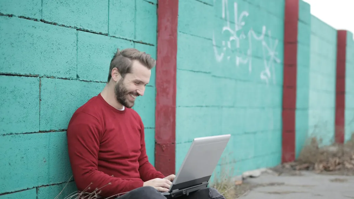 Man in a red sweater using a laptop against a graffiti-covered wall, outdoors, embodying remote work and tech lifestyle.